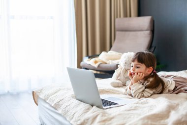 Beautiful little girl lying on a bed in a bedroom and watching something on a laptop
