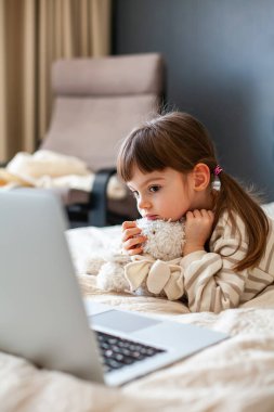 Thoughtful cute little girl hugging her bunny toy lying on a bed in a bedroom and watching something on a laptop