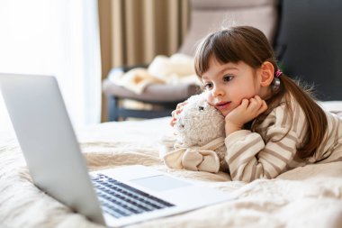 Adorable little girl hugging her bunny toy lying on a belly in a bedroom and watching something on a laptop