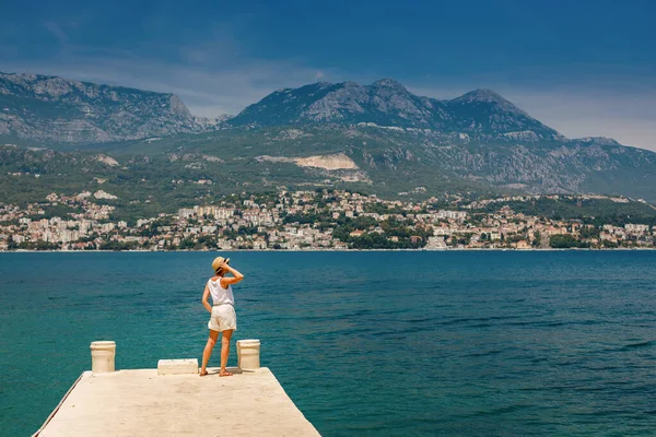 Back view of a woman tourist in white clothes and straw hat standing on the edge of a pier, enjoying beautiful view of Herceg Novi in Montenegro