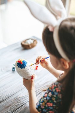 Little girl with bunny ears holding gypsum Easter egg and painting it for Easter decoration