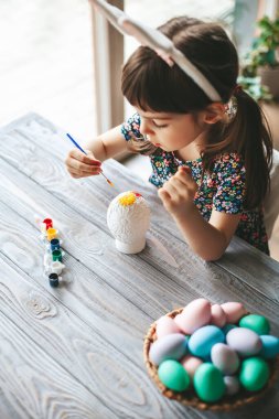Top view little girl with bunny ears sitting at the table painting Easter egg, made of gypsum
