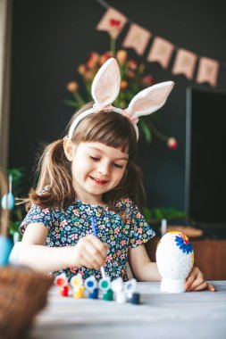 Tavşan kulaklı gülümseyen kız evde oturmuş boya fırçasıyla Paskalya yumurtası boyuyor. Paskalya kutlaması kavramı.