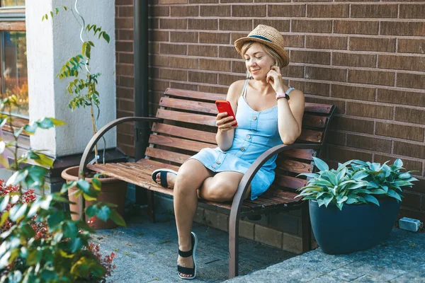 Hasır Şapkalı Gülümseyen Kadın Bahçedeki Bankta Otururken Akıllı Telefon Kullanıyor — Stok fotoğraf