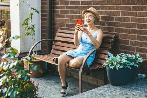 Hasır Şapkalı Gülümseyen Kadın Bahçede Bir Bankta Otururken Telefonunu Kullanıyor — Stok fotoğraf