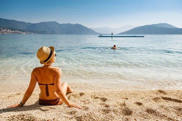 戴着草帽的女人坐在黑山海滩上 看着大海 享受着阳光灿烂的日子 暑假的概念 — 图库照片