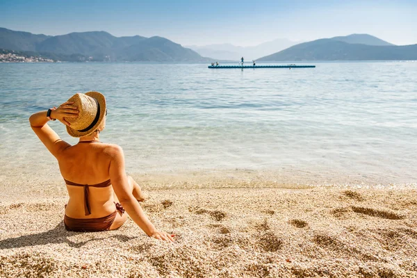 穿着棕色泳衣和草帽的女人坐在黑山海滩上 享受着阳光灿烂的日子 暑假的概念 — 图库照片