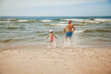 Babası ve küçük kızı denize doğru koşarken eğleniyorlar.