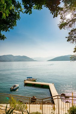 Kotor Körfezi 'nin resimli manzarası. Ağaçların arasından iskeleye ve denizde yüzen eski teknelere bakıyor.