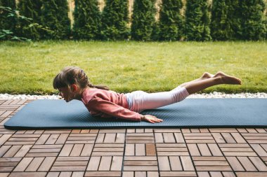 Çekirge pozisyonunda yatan 5-6 yaşlarında küçük bir kız ya da dışarıda yoga yapan bir salabhasana. Çocuklar için yoga. Çocuğun sağlıklı yaşam tarzı..