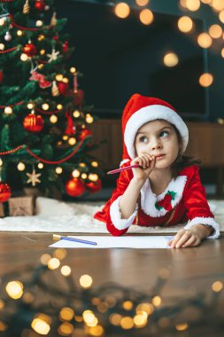 Noel ruhu havadayken, küçük bir kız Noel Baba için dilek listesine odaklandı, arka planda yeni yıl ağacı.