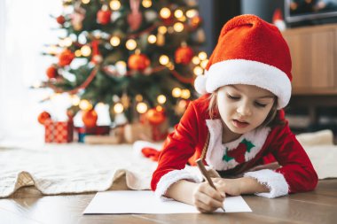 5-6 yaşlarında, Noel Baba 'nın şapkasıyla yeni yıl ağacının yanında uzanmış, Noel Baba' ya yürekten bir mektup yazan tatlı bir kız.