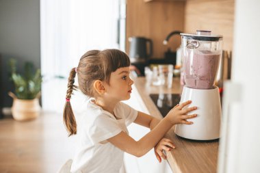 Lezzetli ve besleyici bir içecek yapmak için blender kullanan tatlı küçük kız mutfakta duruyor.