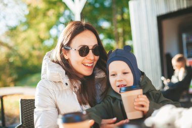 Mutlu kadın ve küçük oğlu sıcak giysiler içinde güneşli açık hava kafesinde sıcak içecekler içiyorlar.