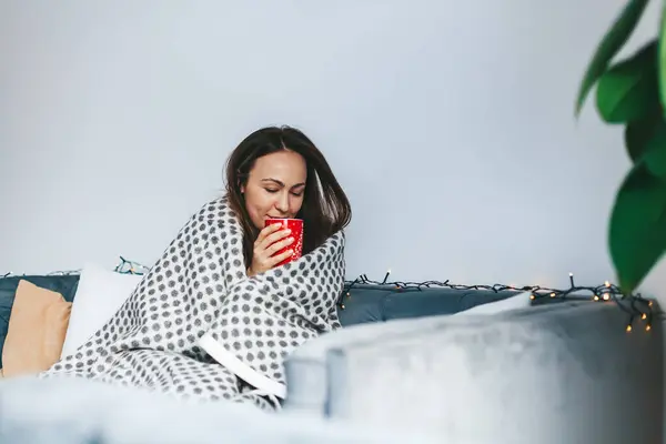 Woman drinking a hot beverage sitting on the couch, wrapped in warm and comfort blanket