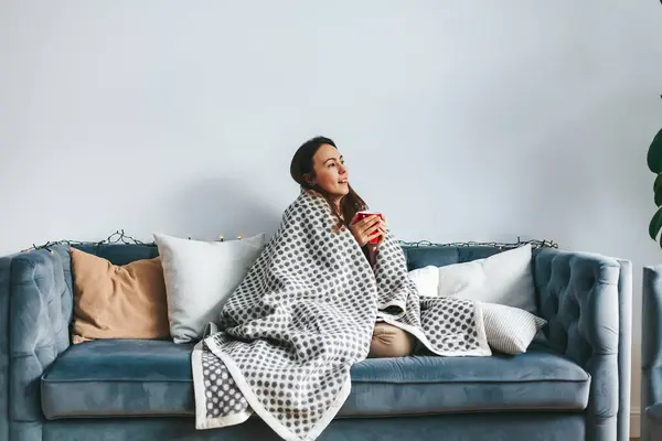 Wrapped in warm and comfort blanket, a woman enjoying a quiet moment on the couch with a cup