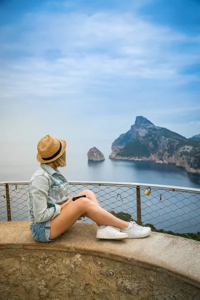 Mirador de El Colomer 'in kenarında oturan hasır şapkalı bir kadın Mallorca, İspanya' daki Kaptan Formentor 'un keyfini çıkarıyor.