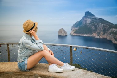 Hasır şapkalı bir turist kadın Mirador de El Colomer 'in kenarında oturuyor, Mallorca, İspanya' daki Kaptan Formentor 'a bakıyor.