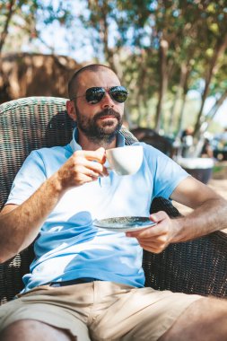 Güneş gözlüklü sakallı bir adam, açık kafede kahve molası veriyor.