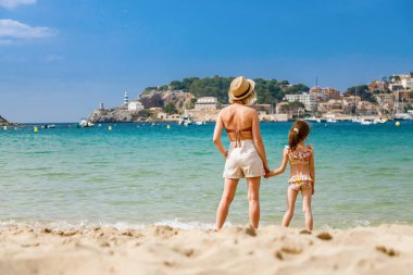 Woman and her little daughter in swimsuit standing together on the beach, watching at the Port de Soller bay in Mallorca. Holidays with children concept. clipart