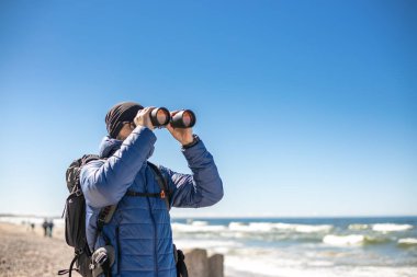Deniz kıyısında duran bir adam, elinde dürbünle, kıyı gözlemiyle meşgul, seyahat deneyimine görsel bir boyut ekliyor.