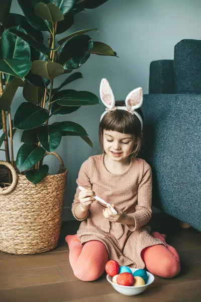 6歳の少女が家で床に座ってイースターエッグを描いている カラフルなイースターエッグが近くのボウルに入っています イースターコンセプト ストックフォト