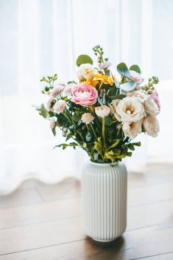 Değişik renkte ve tipte canlı bir çiçek buketi, beyaz, tahta bir zeminde tırtıklı vazoda, pencerenin arka planına karşı, katıksız perdelerle, sahneyi doğal ışıkla aydınlatıyor.