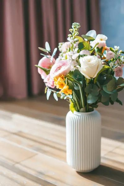 一束鲜花 包括白玫瑰 粉红的兰花和黄色的花朵 装在一个带条纹的白色花瓶里 放在粉红色窗帘的背景下的木制地板上 图库照片