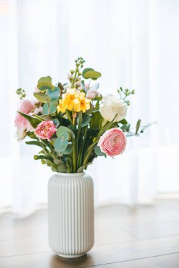 Renk ve tiplerden oluşan canlı bir buket, pencerenin zeminine dayanan, yumuşak doğal ışıkla yıkanmış, beyaz bir vazoda tutulan bir dizi çiçek patlar.