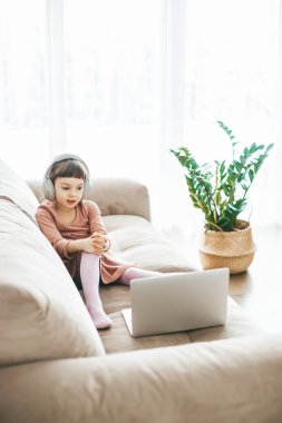 Dizüstü bilgisayar seyrederken kulaklık takan, kanepede oturan 5-6 yaşlarında tatlı bir kız. Kavram: teknoloji ile aşılanmış rahatlama, çevrimiçi eğitim, teknolojik eğlence
