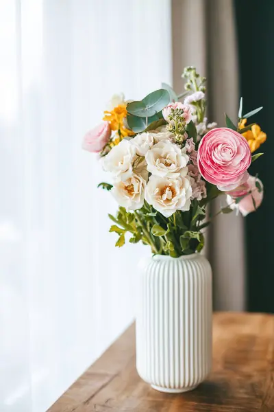 花束一种五彩缤纷的花束 有各种颜色和类型 装在木制表面的一个白色有肋的花瓶中 置于有白色窗帘的窗户的后面 免版税图库图片