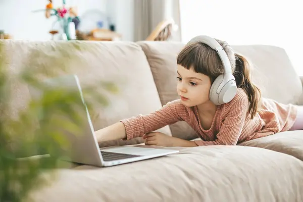 一个迷人的小女孩躺在米黄色的沙发上 戴着耳机 看着笔记本电脑屏幕 技术灌输的放松 在线教育 技术娱乐 图库图片
