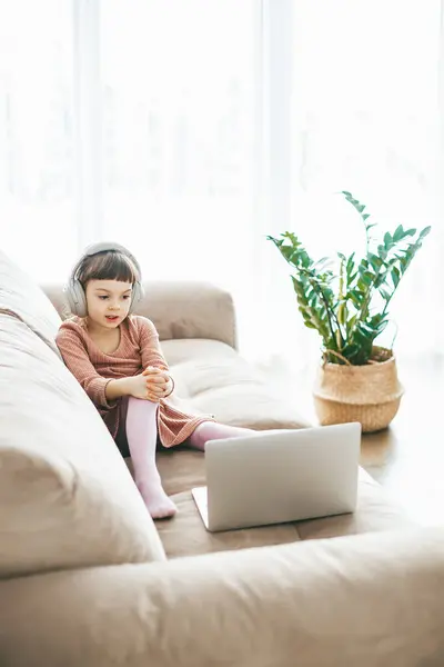 一个甜美的5 6岁女孩坐在沙发上 一边看着笔记本电脑 一边戴着耳机 技术灌输的放松 在线教育 技术娱乐 图库图片