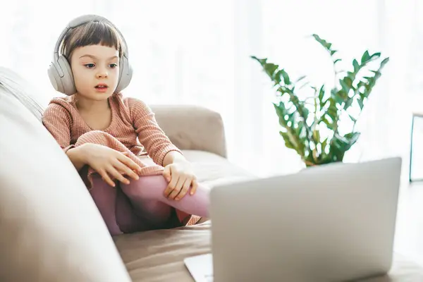 一个坐在沙发上 看着笔记本电脑屏幕的小女孩 耳朵上戴着耳机 迷失在虚拟世界里 技术灌输的放松 在线教育 技术娱乐 免版税图库图片