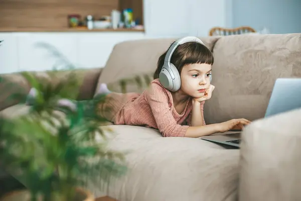 一个小女孩戴着耳机躺在米黄色的沙发上 全神贯注地观看着笔记本电脑屏幕上展现的迷人场景 技术灌输的放松 在线教育 计算机娱乐 免版税图库照片