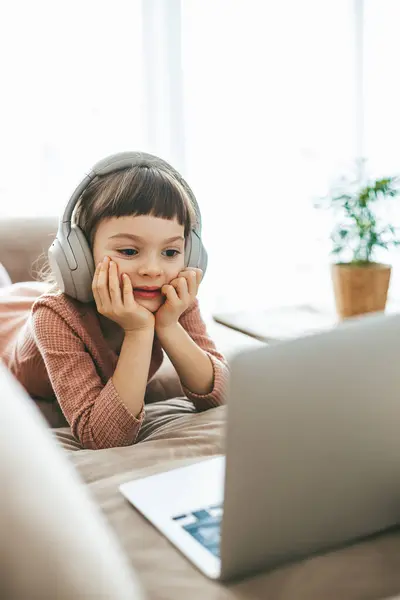 6歳の少女は ソファーでリクライニングしたり ヘッドフォンを着たり ラップトップの画面を見たりする コンセプト テクノロジーを駆使したリラクゼーション オンライン教育 コンピュータエンターテインメント ロイヤリティフリーのストック画像