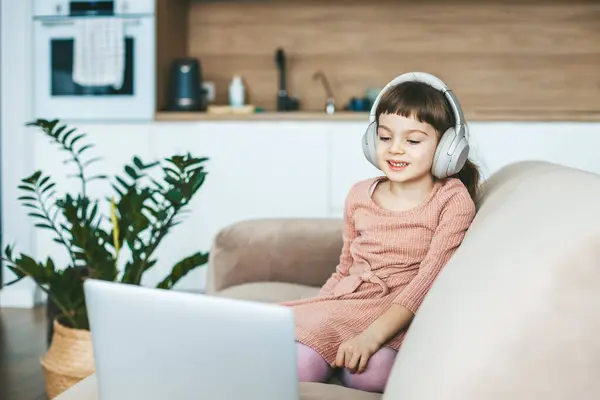 笑顔の5 6歳の少女は ベージュのソファーに座ってラップトップの画面を見ています コンセプト テクノロジーを駆使したリラクゼーション オンライン教育 コンピュータエンターテインメント ロイヤリティフリーのストック画像
