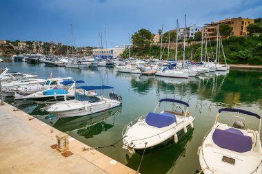 Mallorca 'daki Porto Cristo limanı, kıyı binaları ve yemyeşil alanlar arasında nazikçe demirlemiş teknelerle sakin bir sahne sunuyor.