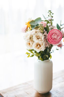 Kaburgalı beyaz bir vazoda sergilenen canlı bir çiçek buketi pencere kenarındaki ahşap bir masa üzerinde duran renk ve çeşitlerden oluşan bir kaleydoskop sergiliyor.