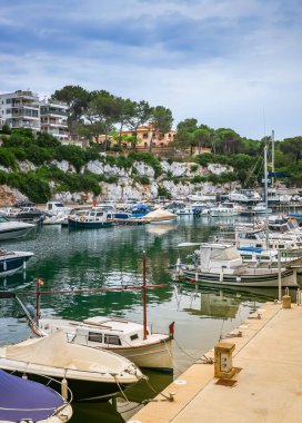 A peaceful atmosphere in Porto Cristo harbor in Mallorca, where moored boats rest calmly amidst the coastal charm of buildings and greenery