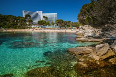 A tranquil scene at Font de Sa Cala beach, Mallorca, showing clear turquoise waters framed by rugged rocks and vibrant greenery clipart