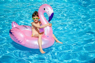 5-6 yaşlarında, pembe bir flamingo şamandırada oturan, çocuklar için yaz eğlencesi ve güvenlik malzemelerini temsil eden genç bir kız.