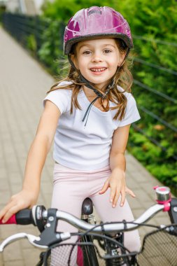 Gülümseyen küçük bir kızın portresi, 5-6 yaşlarında, pembe kasklı ve beyaz tişörtlü, bir bisikletin üzerinde oturuyor.