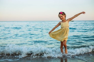 Gülümseyen küçük kız 5-6 yaşlarında sahilde eğleniyor, suda dans ediyor, tasasız ve unutulmaz bir çocukluk deneyimini temsil ediyor.