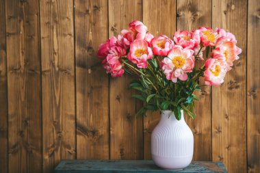 Pembe bir vazoda pembe şakayıklar sergileniyor. Fotokopi alanı olan ahşap bir arkaplanda sergileniyor. Çekici ve zarif bir sahne yaratıyor.