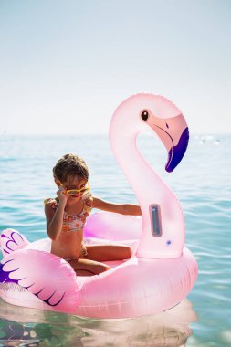 Güneş gözlüklü küçük bir kız pembe bir flamingo üzerinde otururken göz kırpıyor şişirilebilir yüzüyor, denizin tadını çıkarıyor, ılık havanın tasasız bir anını temsil ediyor.