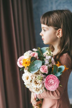 Güzel küçük kız elinde bir buket çiçekle pencerenin yanında duruyor, pembe, beyaz ve sarı çiçekleri birleştiriyor, güller ve ranunculus da dahil.