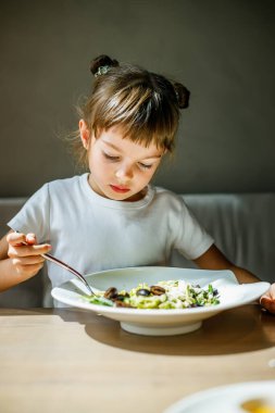 5-6 yaşlarında küçük bir kız, yeşil soslu ve siyah zeytinli vejetaryen makarna yemeğinin tadını çıkarırken yemek masasında oturuyor.