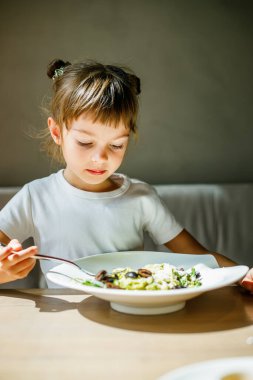 Küçük bir çocuk, 5-6 yaşlarında, yeşil soslu vejetaryen makarna yiyor ve masada siyah zeytin yiyor, sevimli ve memnun görünüyor.