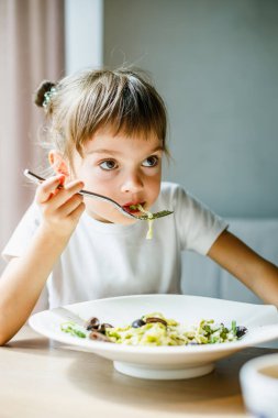 5-6 yaşlarında, yeşil soslu ve siyah zeytinli vejetaryen makarnanın tadını çıkaran genç bir kız.